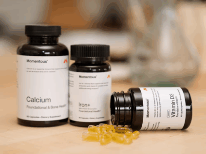 Three bottles of Momentous dietary supplements labeled Calcium, Iron+, and Vitamin D3 sit on a table, with several yellow capsules spilled in front of the Vitamin D3 bottle.