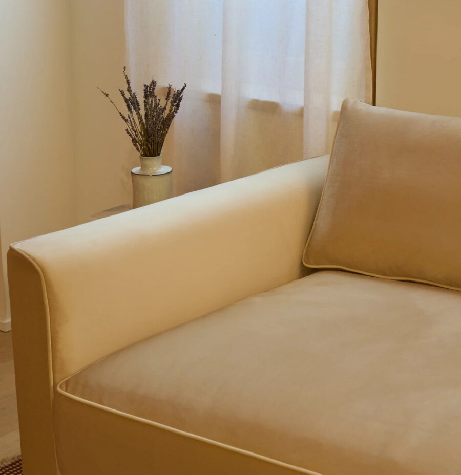 A beige sofa with a matching cushion next to a small table holding a vase of dried flowers, set against a window with sheer curtains.