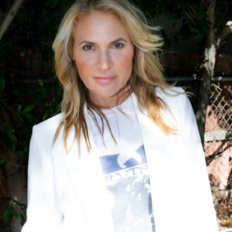 A woman with blonde hair wearing a white blazer and a printed T-shirt stands outdoors in partial sunlight near trees and a chain-link fence.
