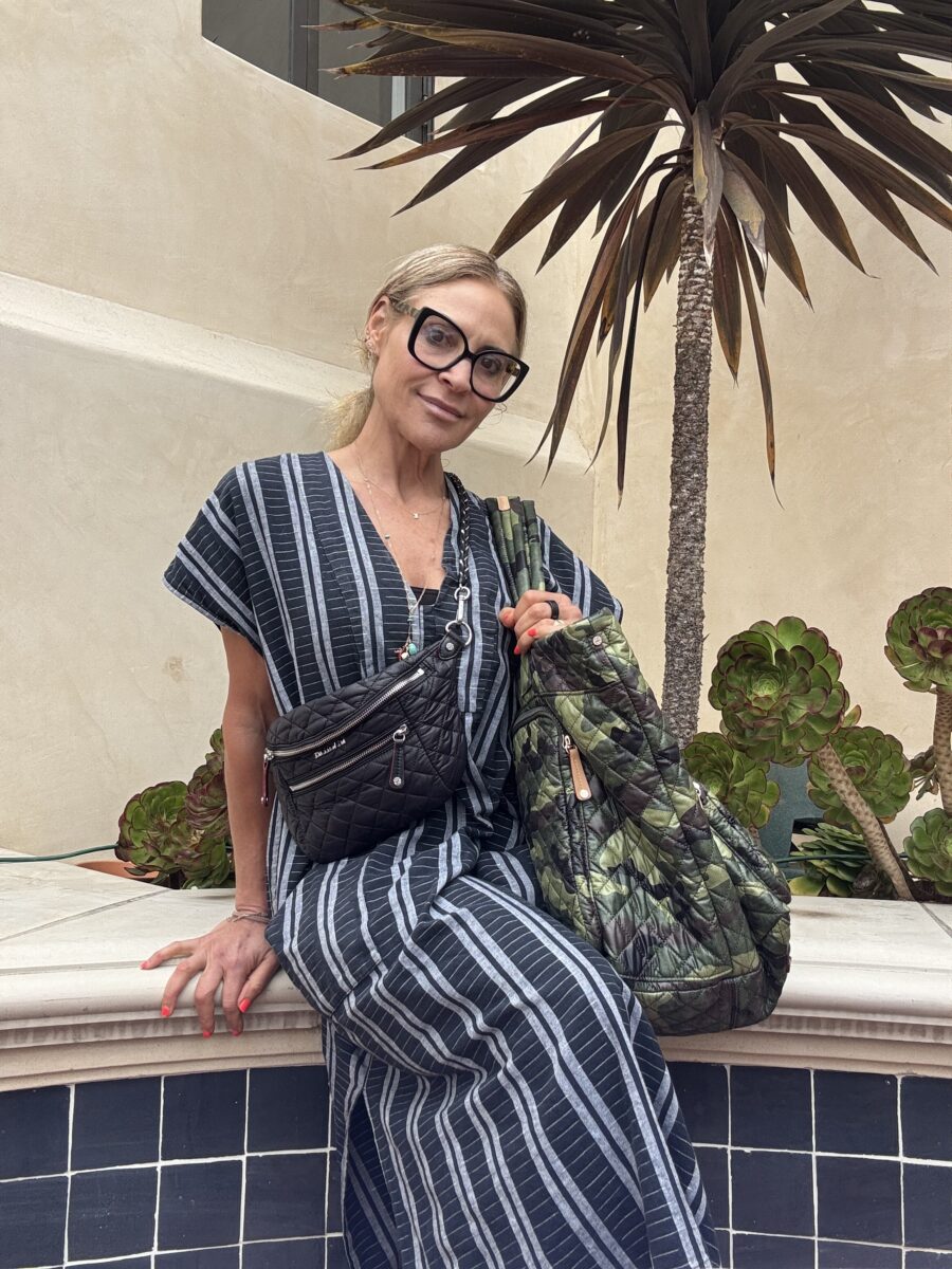 A woman in a striped dress sits on a tiled ledge, holding two quilted bags, with succulents and a tall plant in the background.