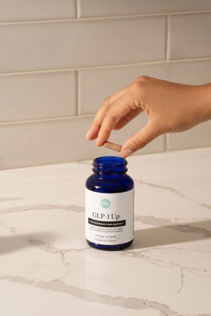 A hand holds a capsule above an open blue bottle labeled "GLP-1 Up" on a marble countertop with a white tile background.