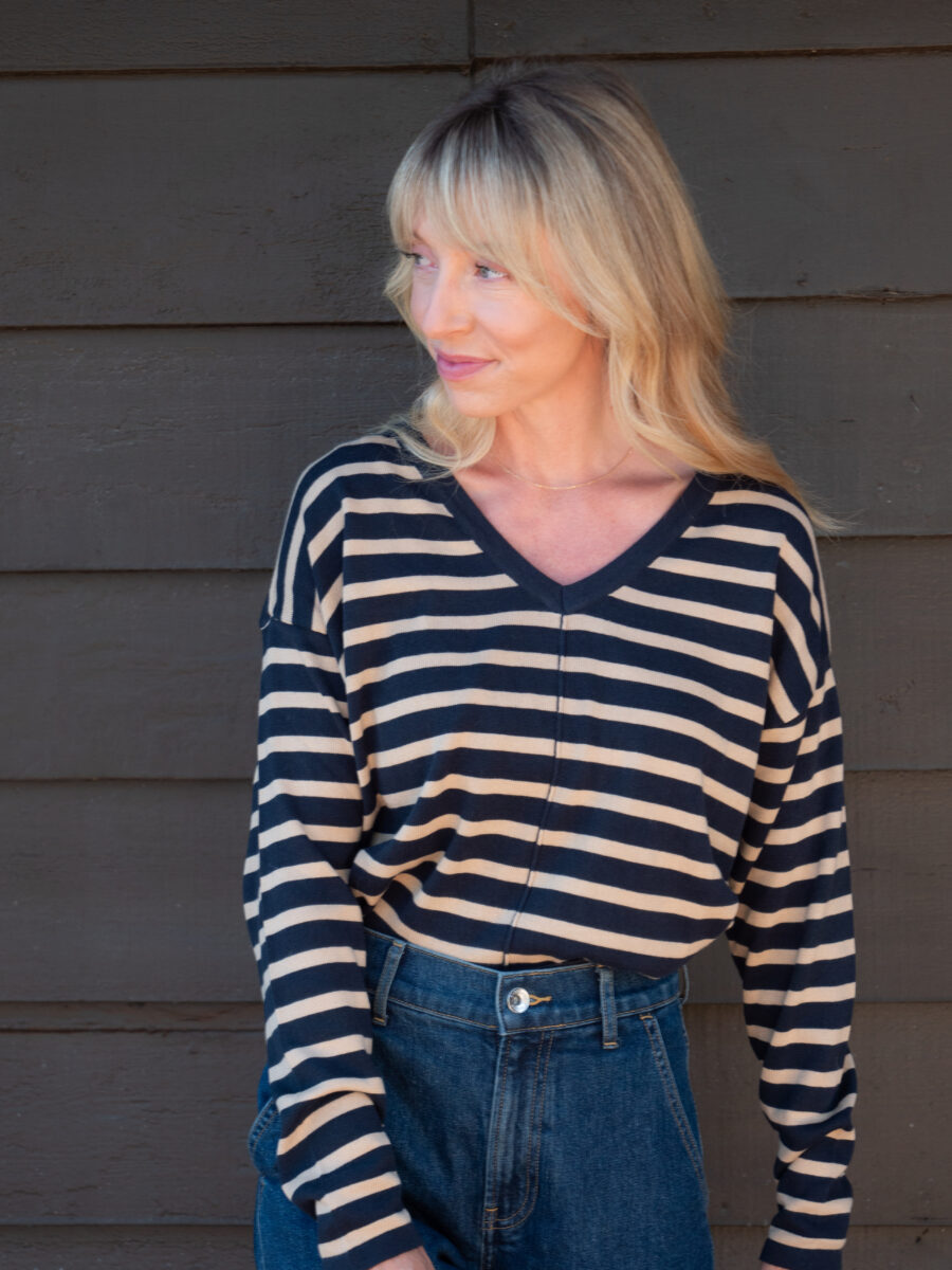 A woman with blonde hair wearing a black and beige striped long-sleeve shirt and high-waisted blue jeans stands in front of a dark wooden wall, looking to the side.