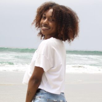 A person with curly hair, wearing a white t-shirt and denim shorts, stands on a beach with waves in the background, smiling at the camera.