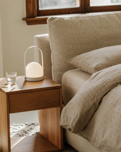 A wooden bedside table with a glass of water and a modern lamp beside a bed with beige linens in a softly lit room.