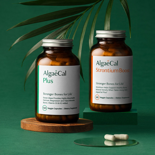 Two bottles of AlgaeCal supplements, AlgaeCal Plus and AlgaeCal Strontium Boost, are displayed on wooden stands with green capsules and a palm leaf in the background.