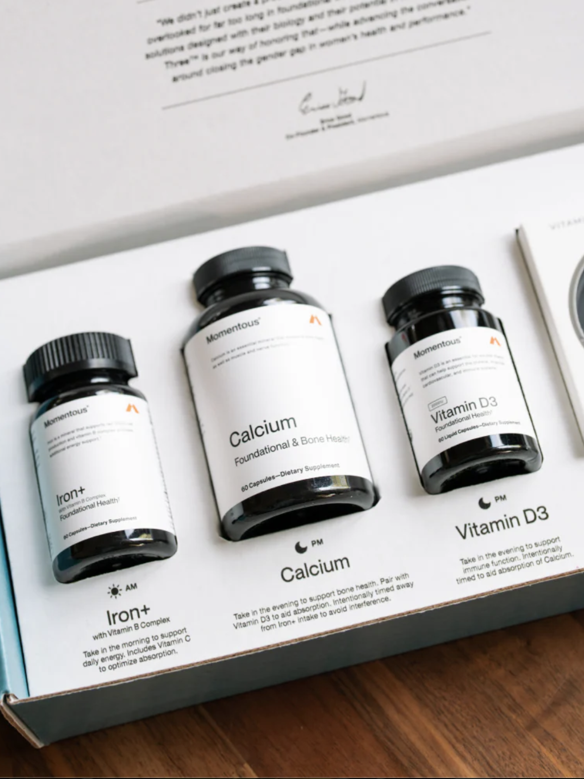 Three supplement bottles labeled Iron+, Calcium, and Vitamin D3 are arranged in a box on a wooden surface, with informational text printed below each bottle.
