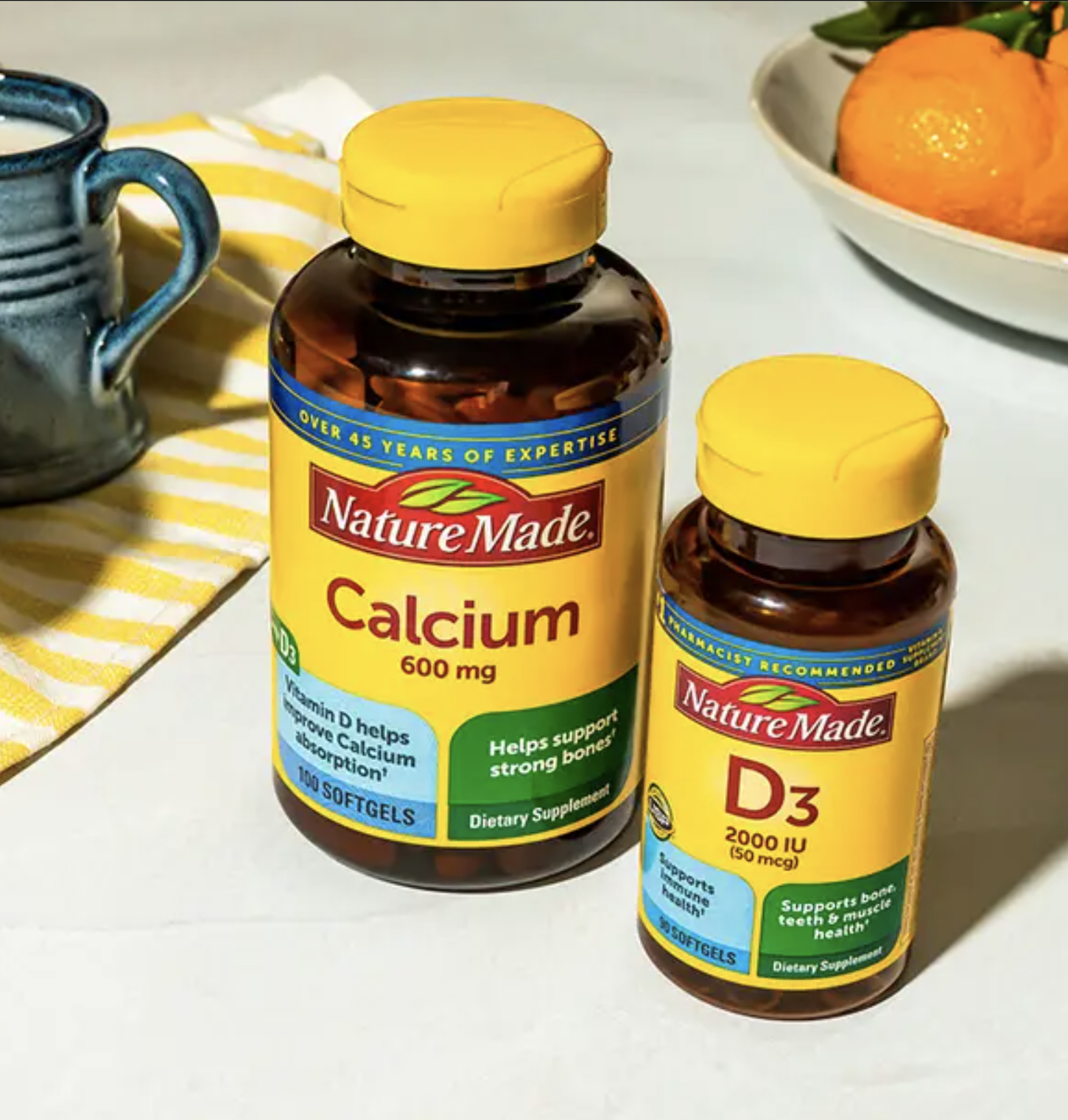 Two bottles of Nature Made supplements, one labeled Calcium 600 mg and the other labeled D3 2000 IU, are placed on a kitchen counter near a blue mug and a plate of oranges.