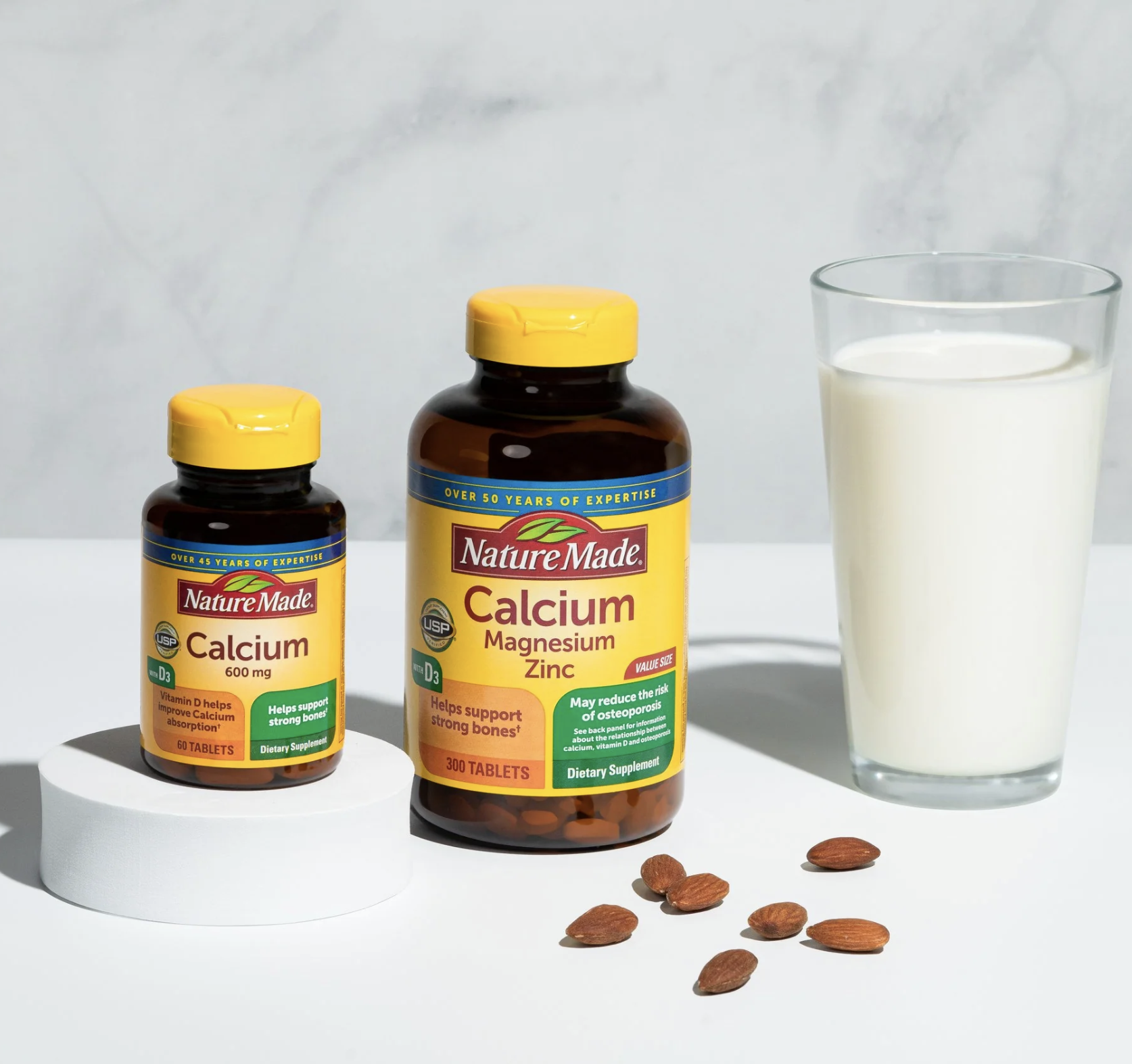 Two bottles of Nature Made calcium supplements, a glass of milk, and several almonds are arranged on a white surface against a light background.