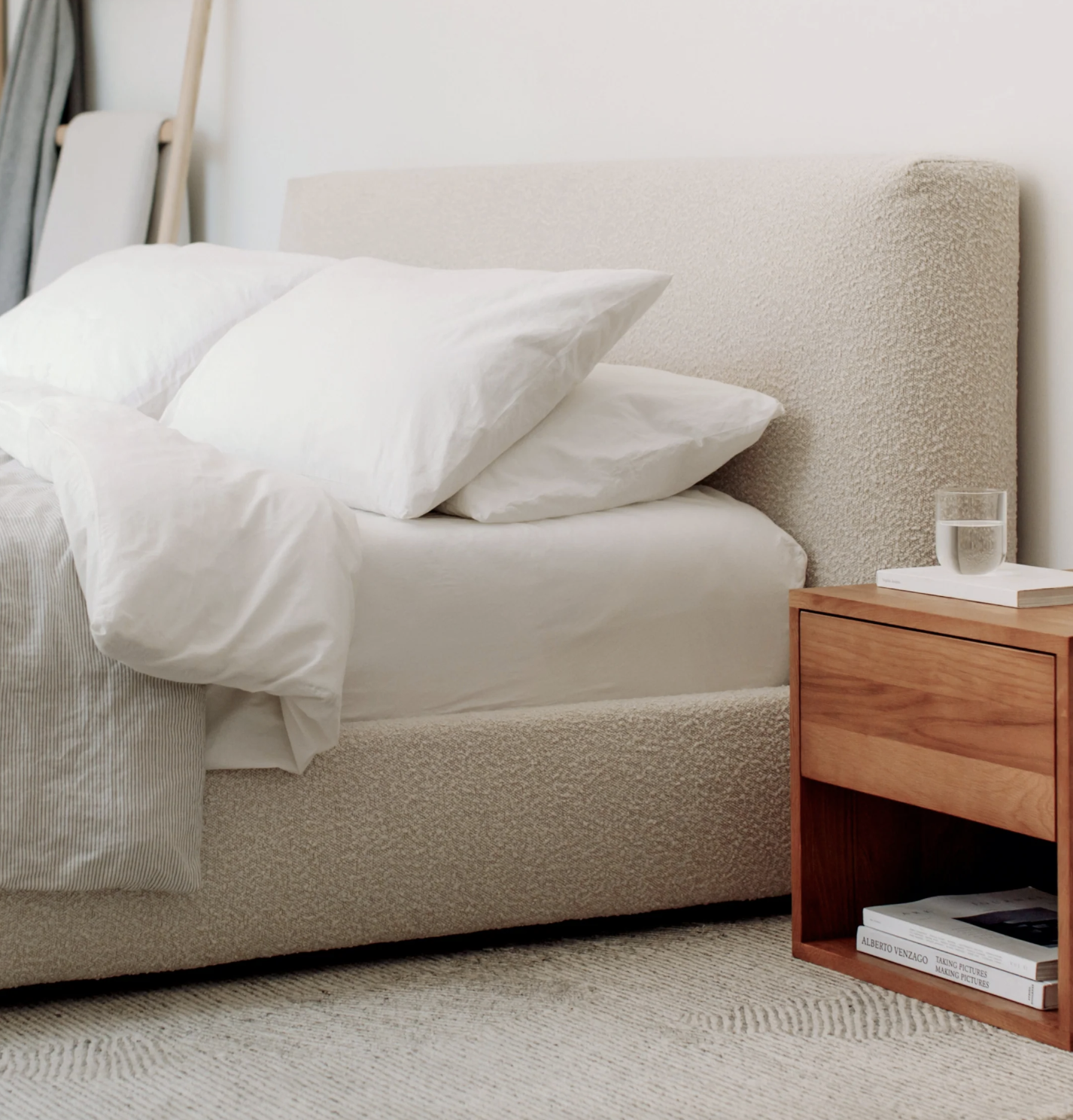 A neatly made bed with white bedding next to a wooden nightstand holding a glass of water and two books, set on a textured rug.