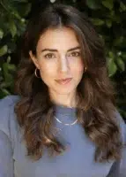 Woman with long wavy brown hair wearing a blue top, gold hoop earrings, and a gold necklace, standing in front of leafy green foliage.
