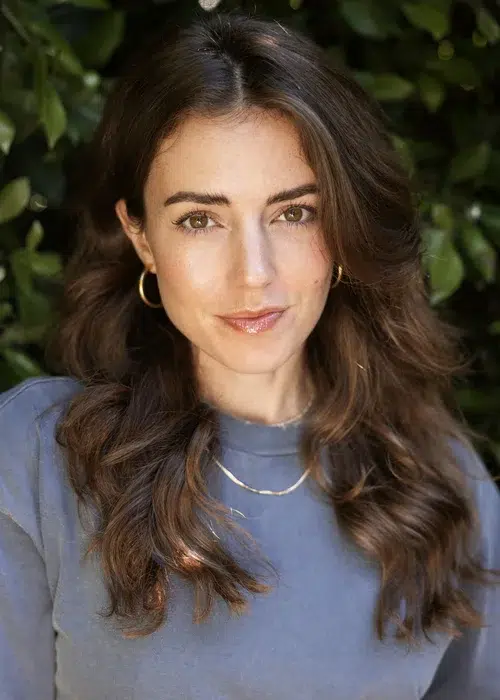 Woman with long wavy brown hair wearing a blue top, gold hoop earrings, and a gold necklace, standing in front of leafy green foliage.
