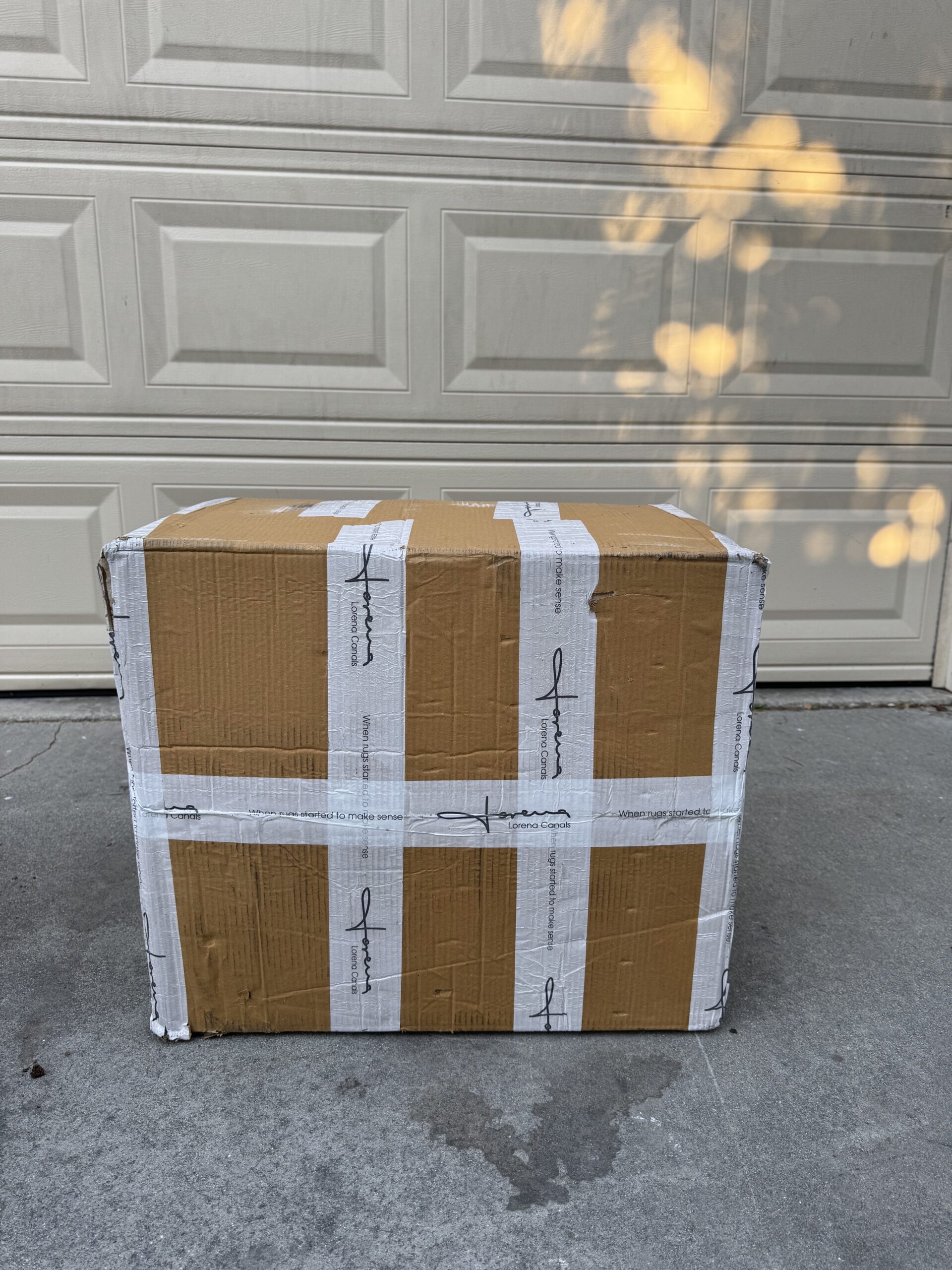 A large, sealed brown cardboard box with white tape sits on a concrete driveway in front of a closed garage door.