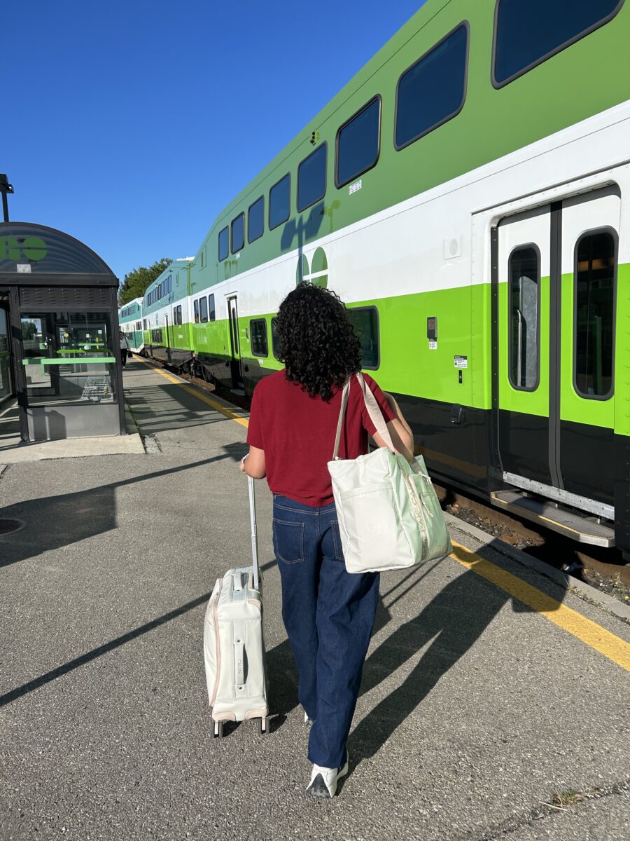 Person with a suitcase and tote bag walks toward a green and white double-decker train at a station on a sunny day.