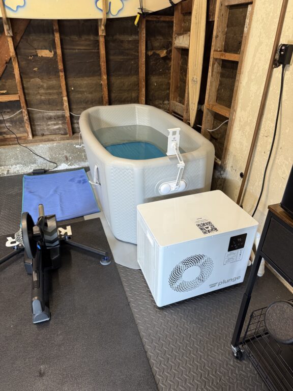 Inflatable ice bath tub filled with water, connected to a white Plunge chiller unit, in a garage with exposed wall studs and exercise equipment nearby.