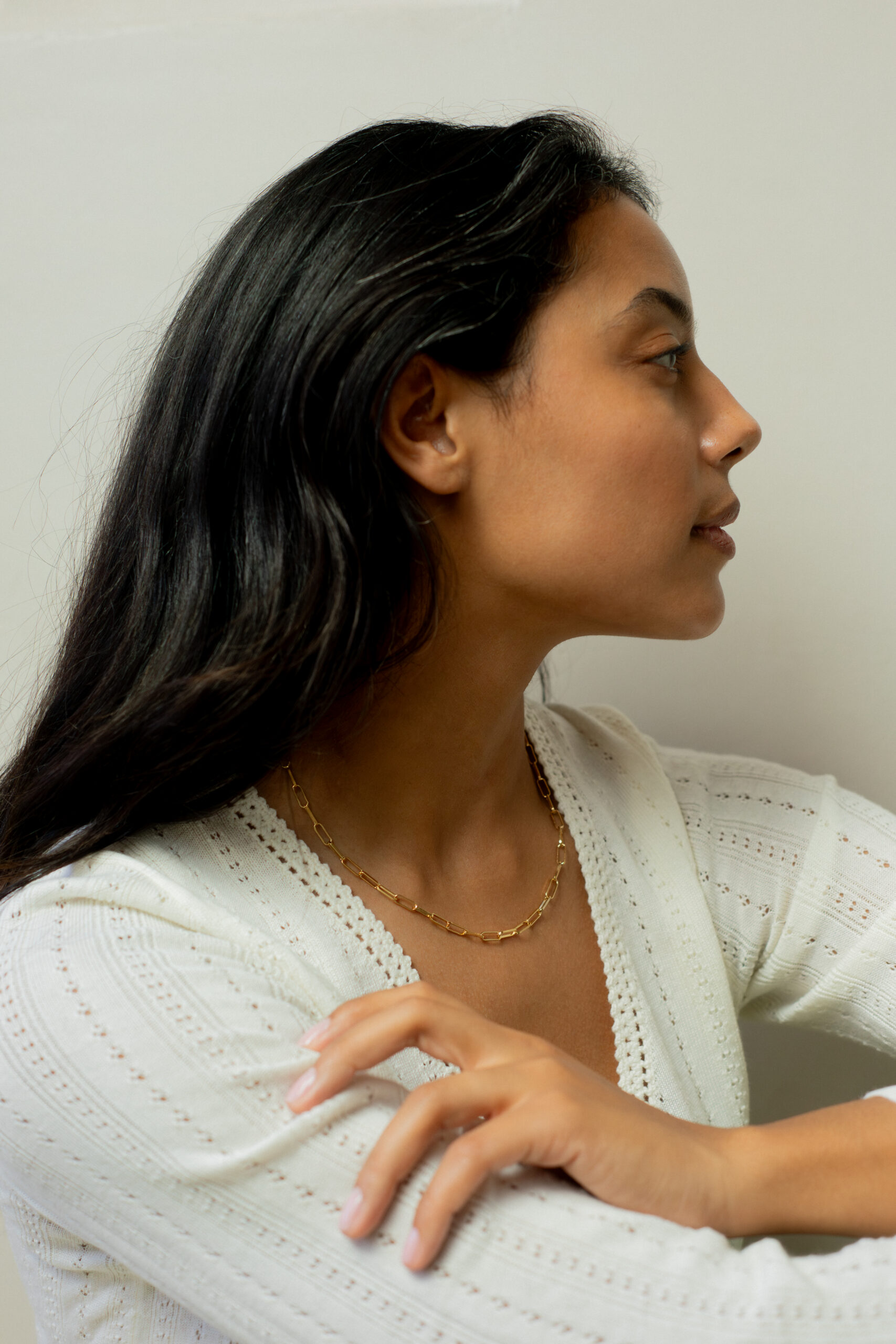 Woman with long dark hair in a white top and gold necklace sits with her arms crossed, looking to the side against a plain background.