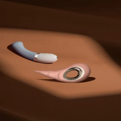 Two modern sex toys, one blue and curved with a silver accent and one pink with a circular opening, placed on a brown, softly lit surface.