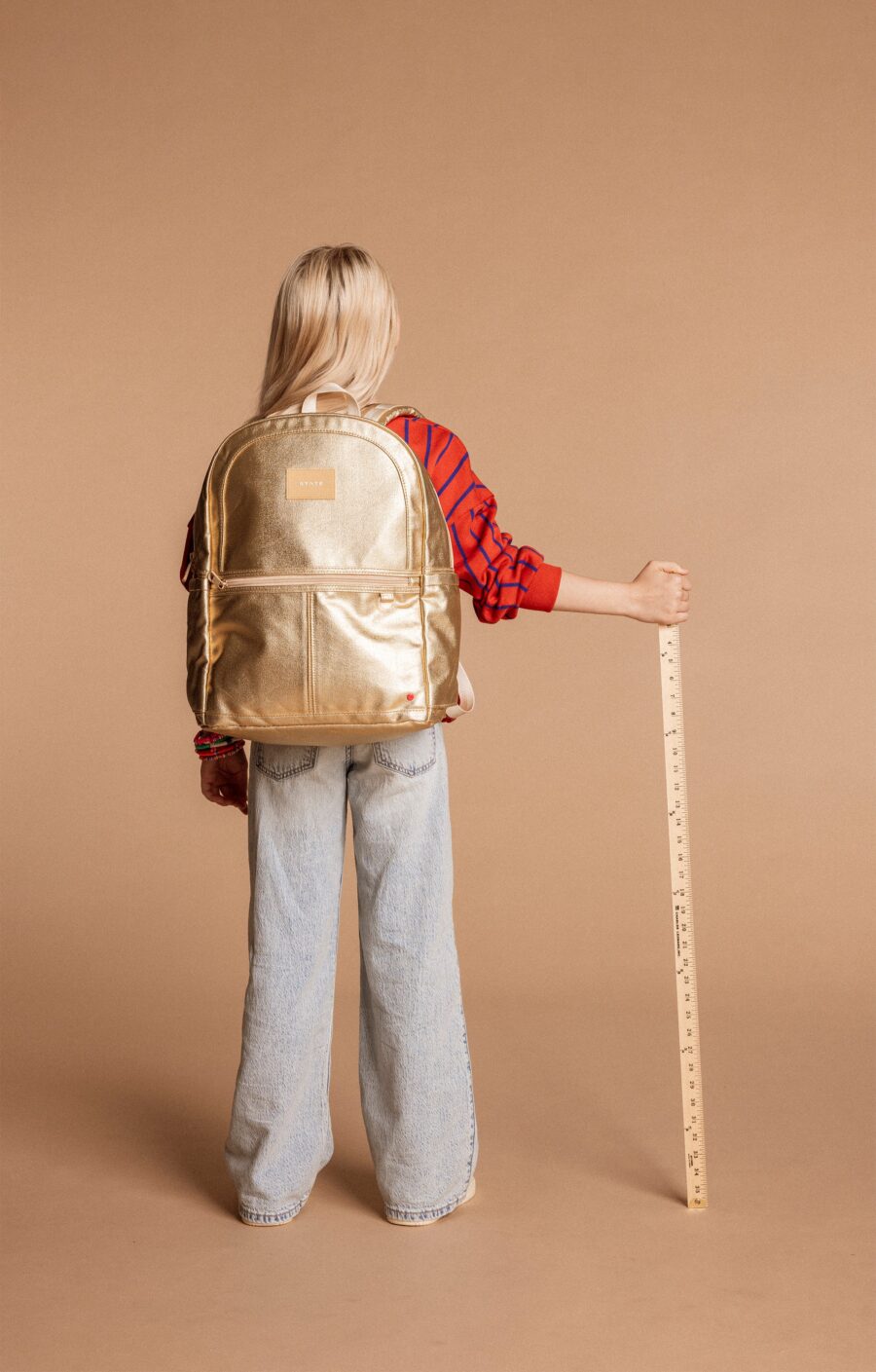 A person with long blonde hair wearing a red shirt and blue jeans stands facing away, holding a yardstick and carrying a large gold backpack.