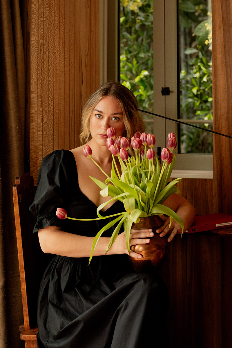 A woman in a black dress sits indoors, holding a brown vase filled with pink tulips, with a window and greenery in the background.