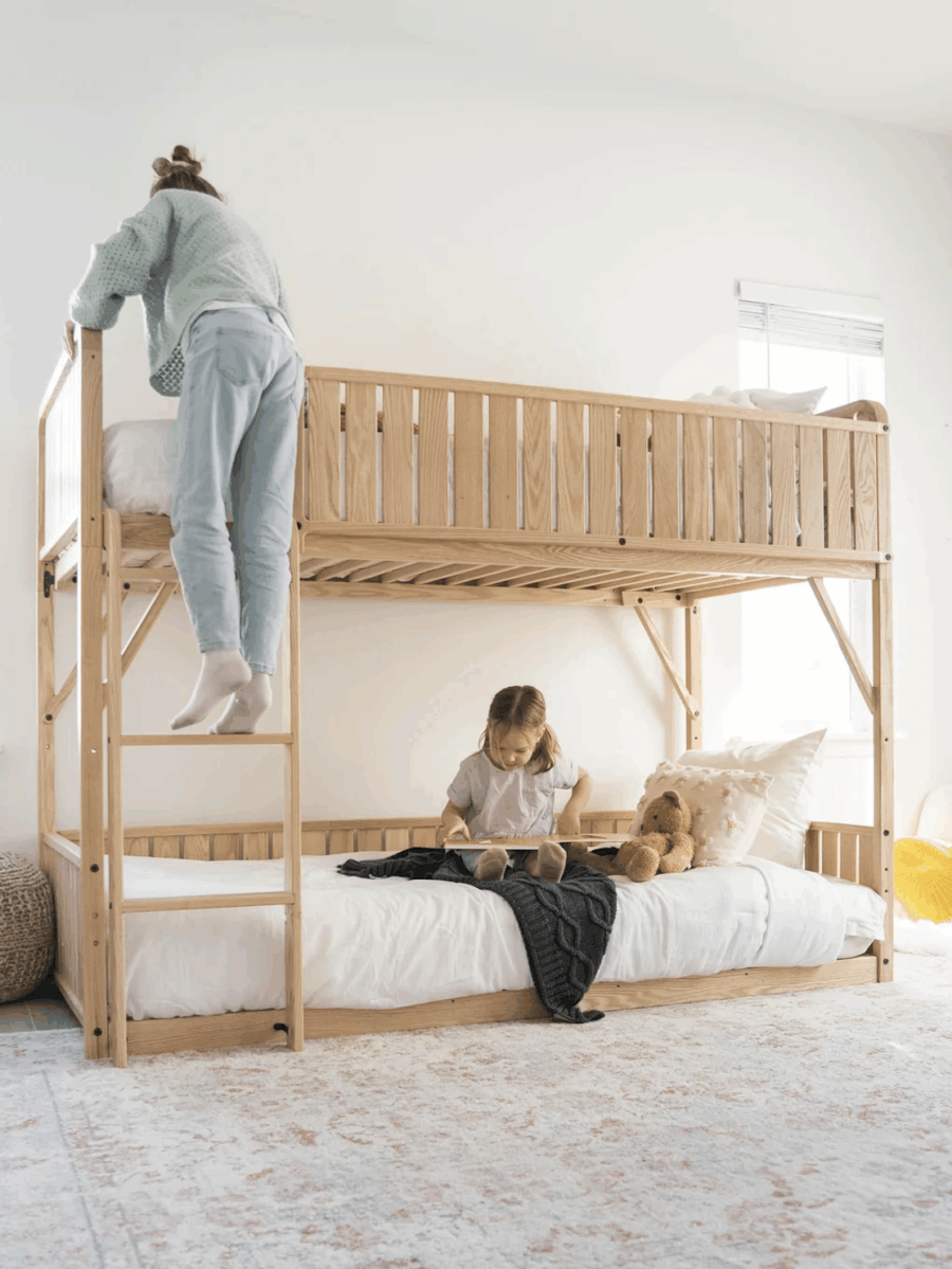 Two children use a wooden bunk bed; one climbs the ladder to the top bunk while the other sits on the lower bunk, reading a book next to a teddy bear.