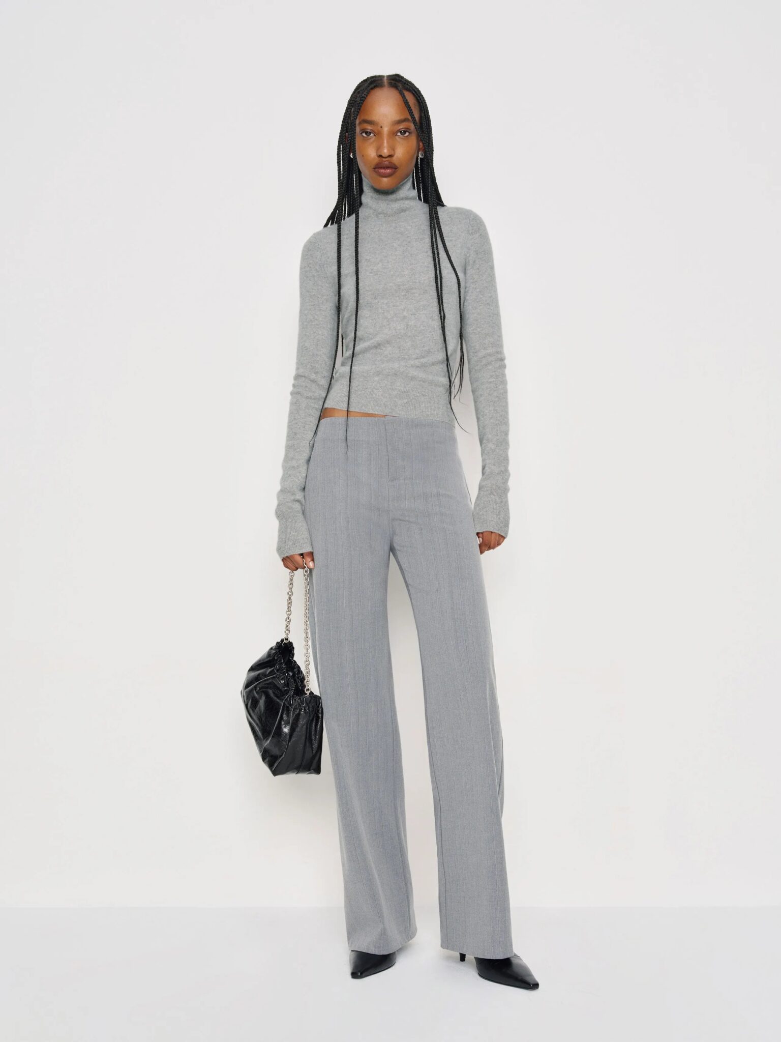A woman wearing a light gray turtleneck sweater, gray high-waisted trousers, and black pointed-toe shoes holds a black handbag against a plain white background.