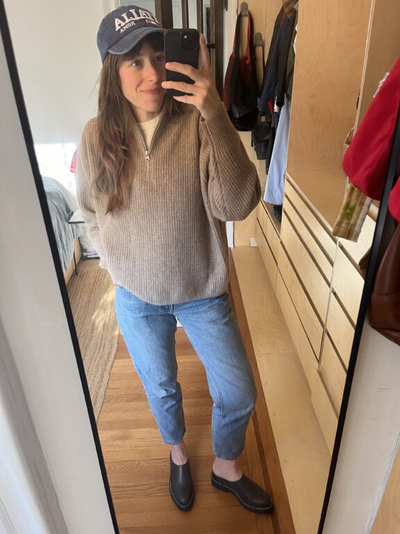 Person stands in front of a mirror taking a selfie, wearing a blue baseball cap, tan sweater, blue jeans, and black slip-on shoes in a tidy room with wooden floors and built-in drawers.