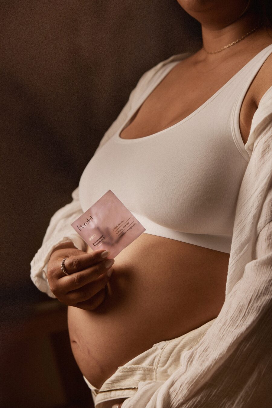 A person wearing a white bralette and open shirt holds a packet labeled "Patch"—a convenient prenatal vitamin—against their bare stomach.