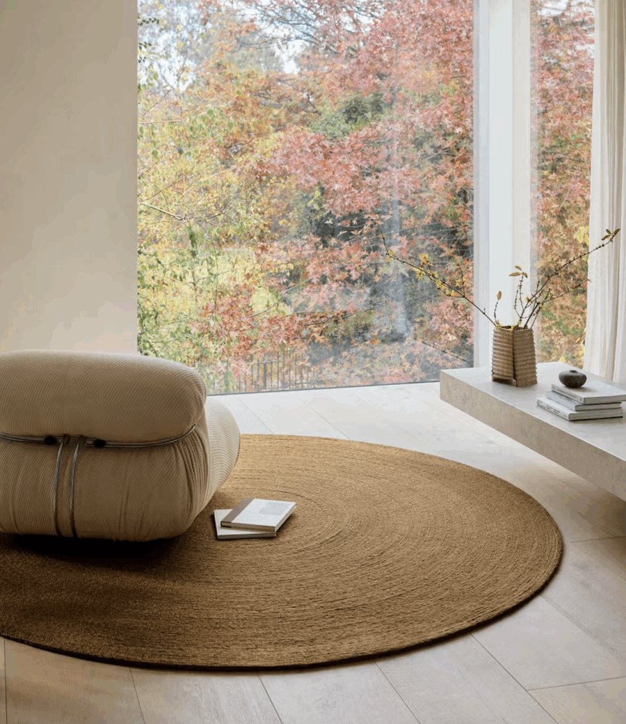 A beige lounge chair and a round woven rug are placed near a large window with a view of autumn trees. A low shelf holds a vase with branches, a bowl, and a stack of books.