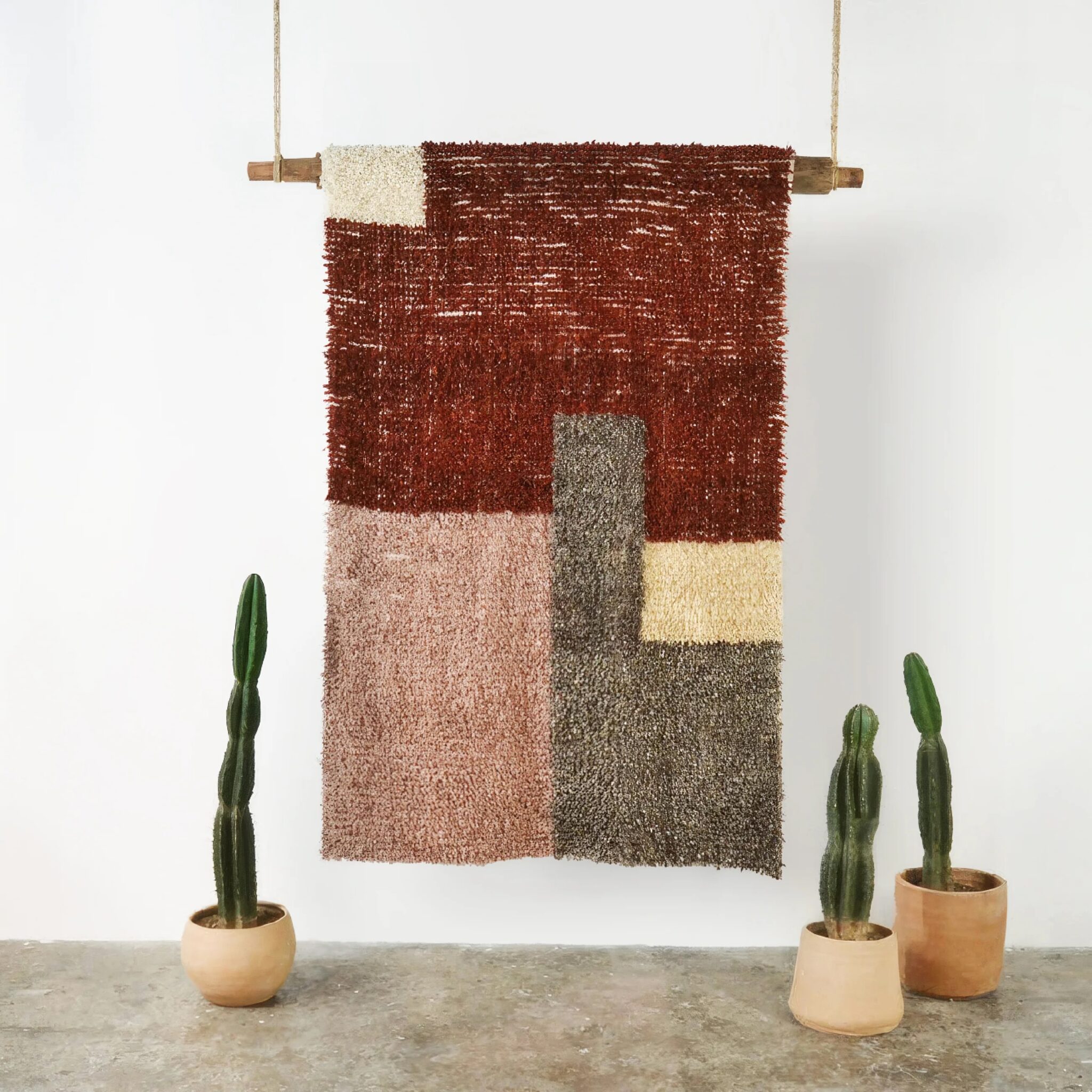 A textured, geometric wall hanging with blocks of brown, beige, and gray tones is suspended on a rod between three potted cacti on a concrete floor.