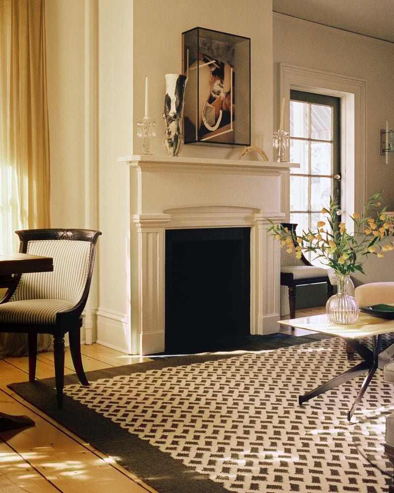 A living room with a black fireplace, a patterned rug, a vase with yellow branches on a coffee table, and framed art above the mantel. Sunlight streams through tall windows with sheer curtains.