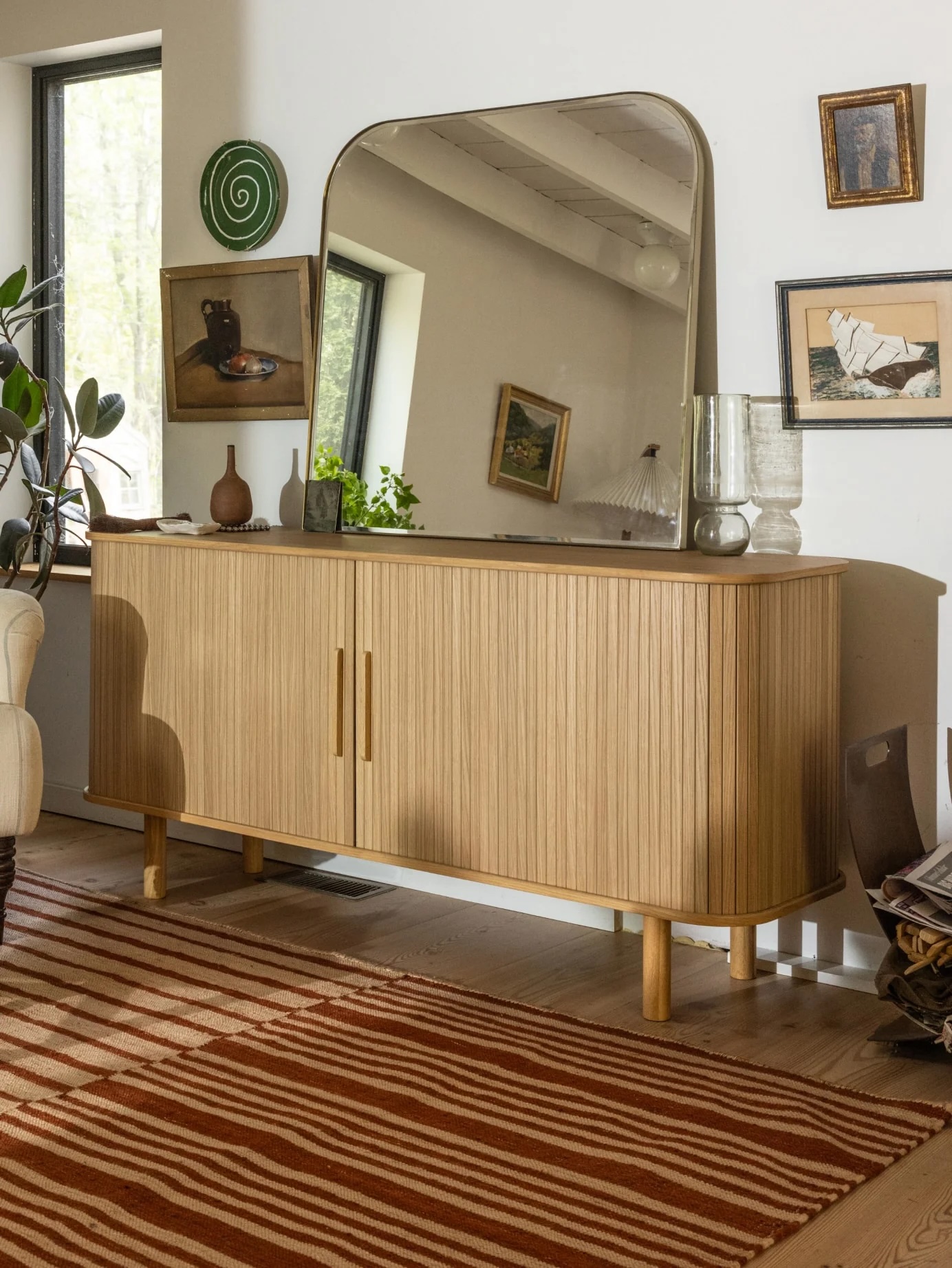 A wooden sideboard with vertical grooves stands against a white wall, topped with a large mirror, vases, and decor. Artwork and a plant are nearby; a striped rug covers the floor.