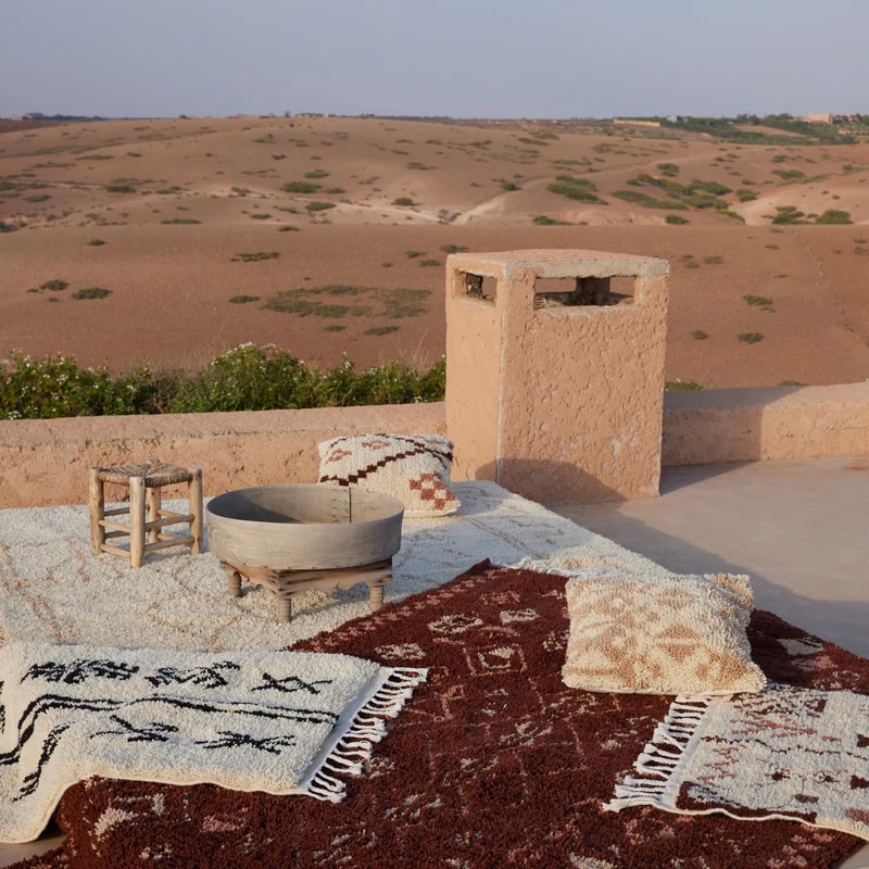 Outdoor patio with patterned rugs, pillows, a small wooden stool, and a round wooden basin, overlooking a desert landscape with sparse greenery.