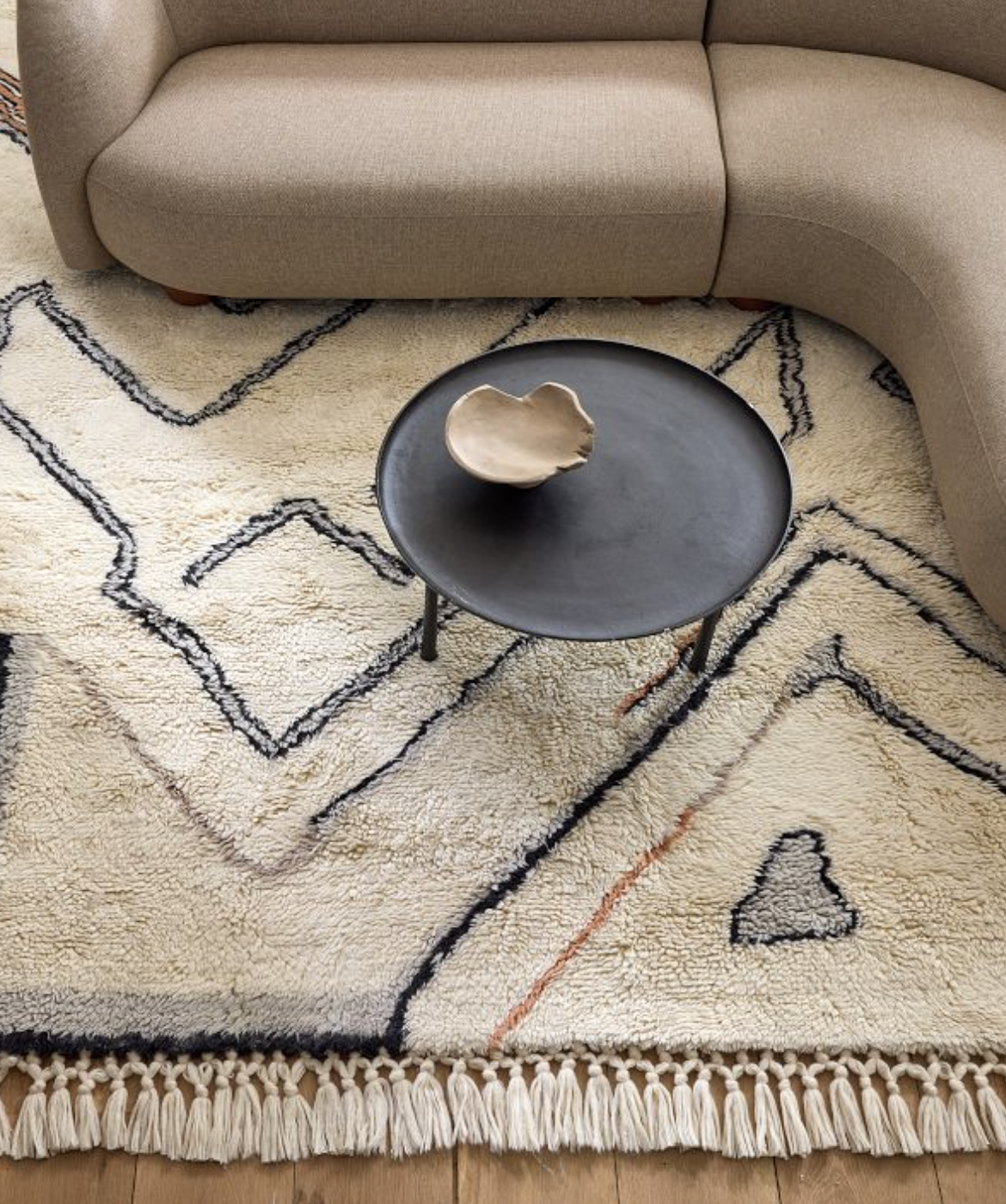 A curved beige sofa sits on a patterned cream rug with black lines, next to a round black coffee table holding a beige ceramic bowl.