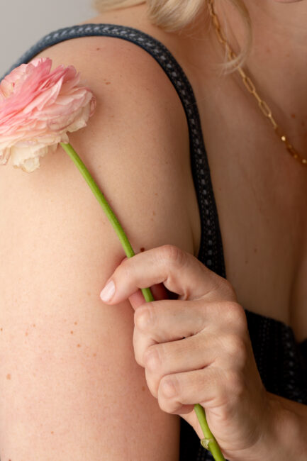 A person holds a pink ranunculus flower against their bare shoulder, wearing a dark sleeveless top and a gold chain necklace.