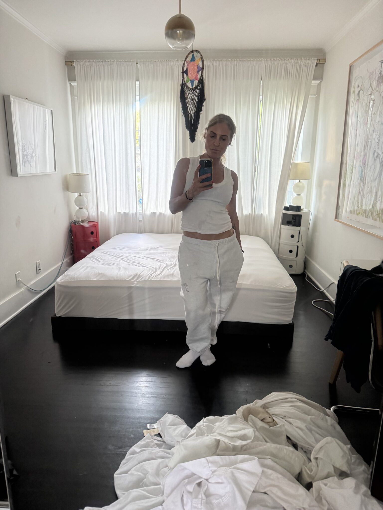 A person in white clothes takes a mirror selfie in a bedroom with an unmade bed, white curtains, and clothes on the floor.