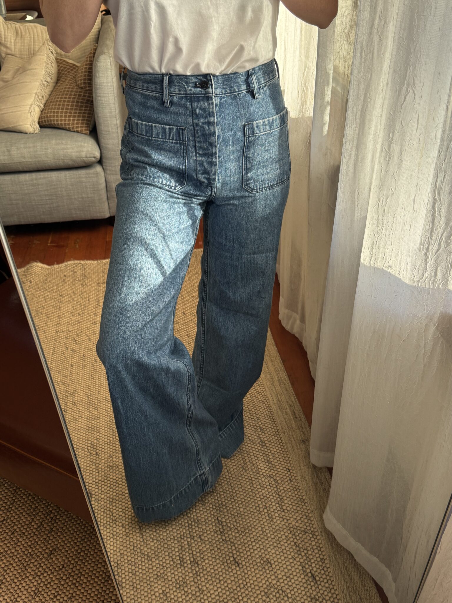 Person wearing high-waisted, wide-leg blue jeans and a white T-shirt, standing indoors on a woven rug in front of a mirror.