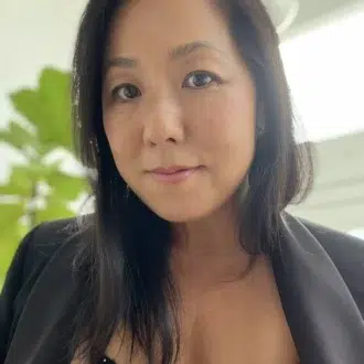 A woman with long dark hair wearing a black blazer and a black polka dot top stands indoors with a green plant and bright window in the background.