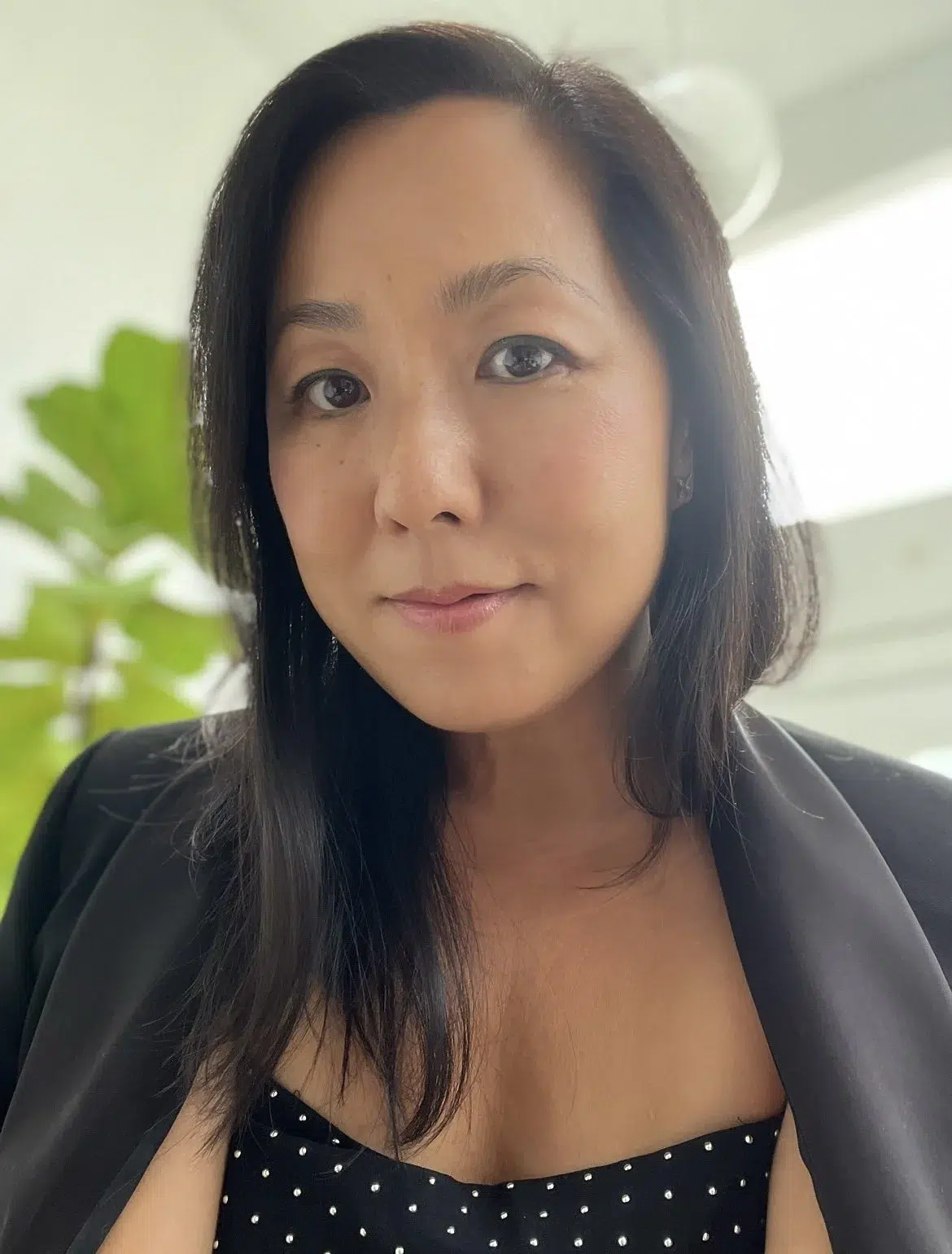 A woman with long dark hair wearing a black blazer and a black polka dot top stands indoors with a green plant and bright window in the background.