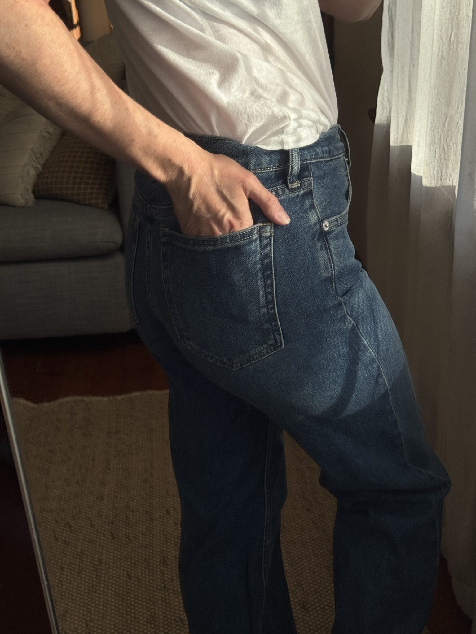 A person wearing a white t-shirt and blue jeans stands near a window with one hand in their back pocket.