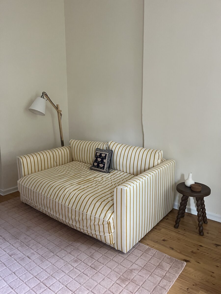 A yellow and white striped sofa with a patterned cushion sits next to a wooden side table and floor lamp on a pink rug in a room with light-colored walls and wooden floor.
