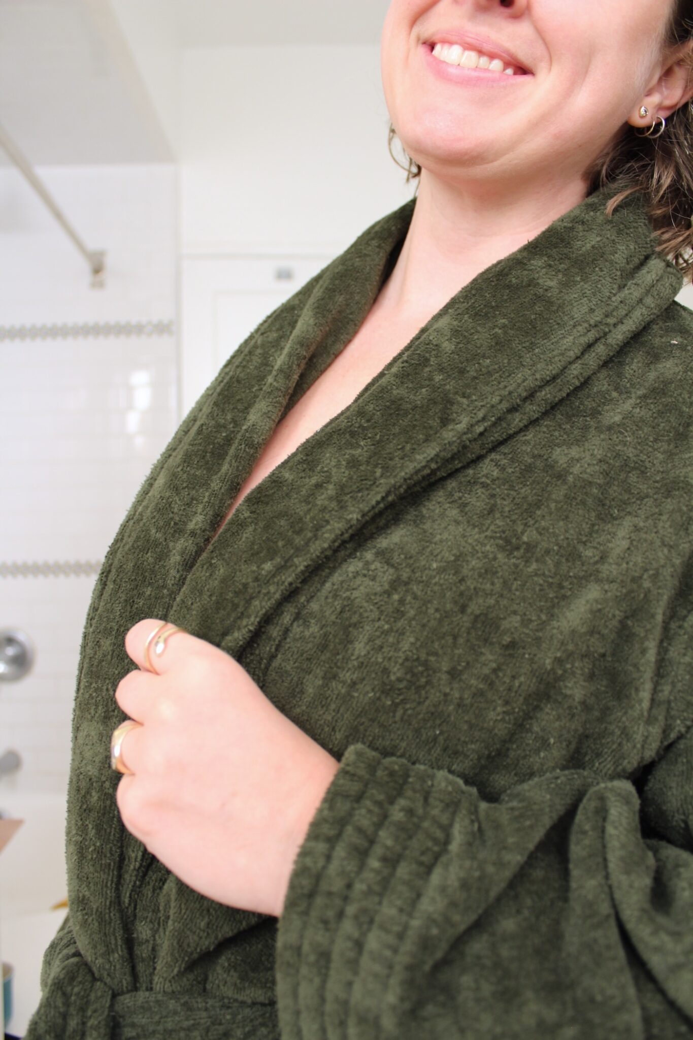 A person wearing a green bathrobe stands in a bathroom, smiling slightly, with white tiled walls and a shower in the background.