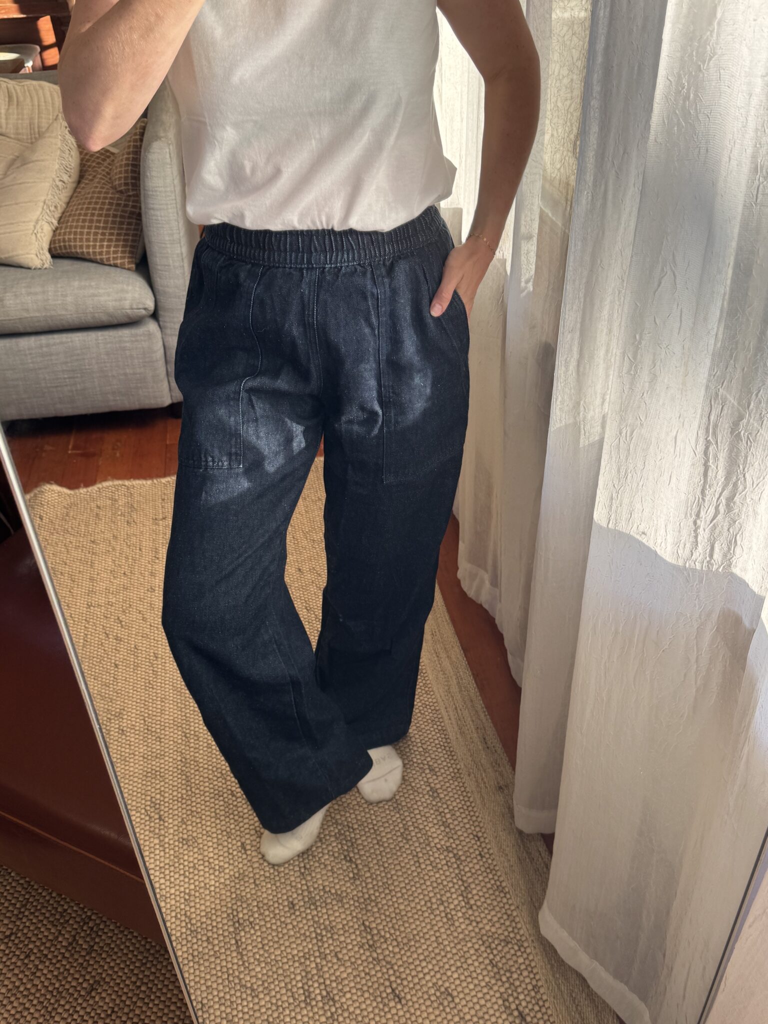 Person wearing a white short-sleeve top, dark blue loose pants, and white socks stands indoors, posing in front of a mirror near a window with sheer curtains.