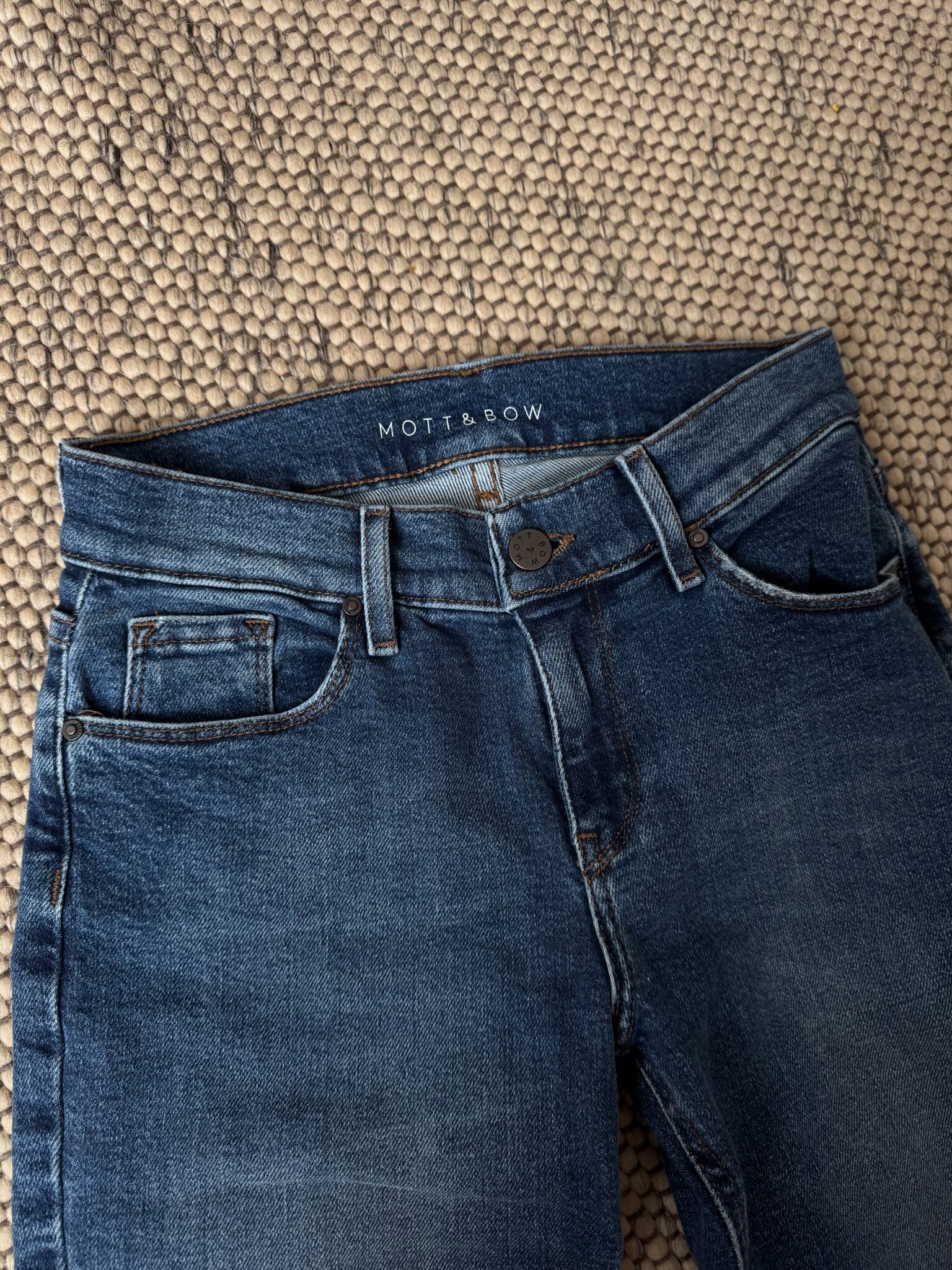 A pair of blue jeans with a "Mott & Bow" label is laid flat on a textured beige carpet.