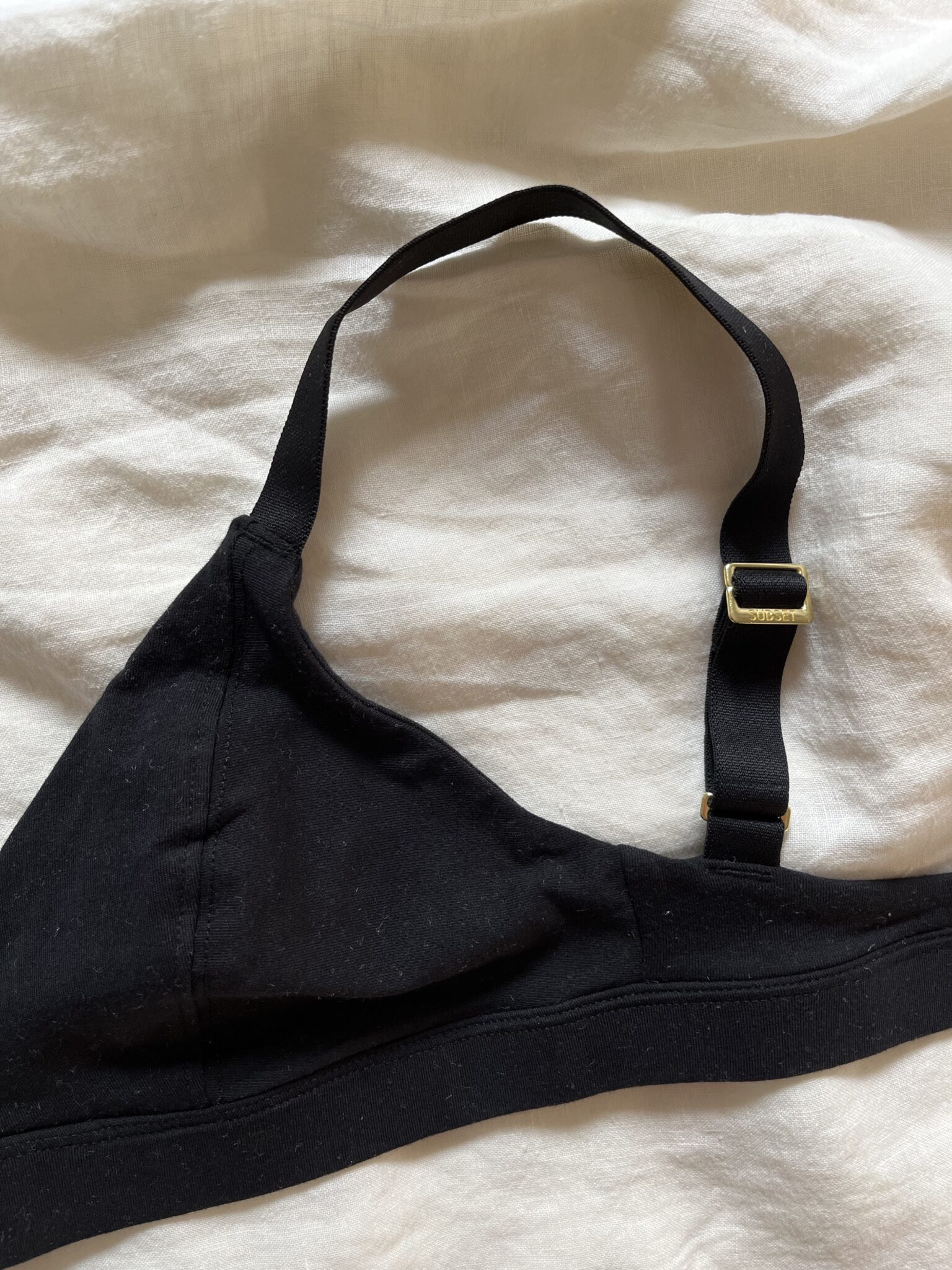 A black bralette with adjustable shoulder straps and gold hardware lies on a cream-colored fabric background.