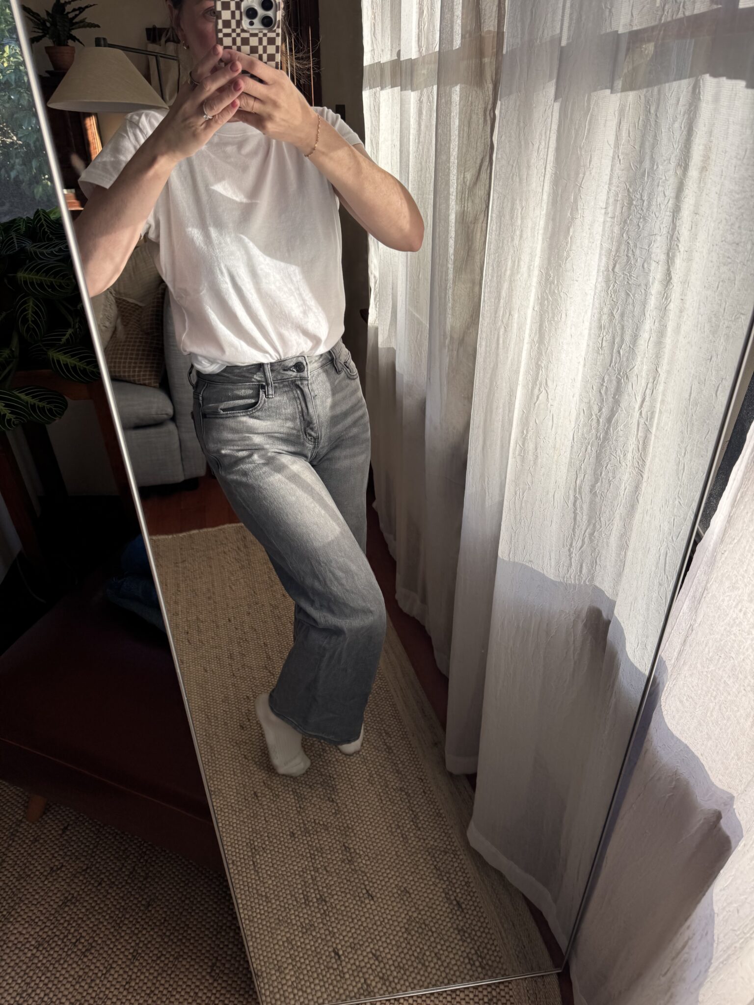 Person taking a mirror selfie wearing a white t-shirt, gray jeans, and white socks, standing in a sunlit room with sheer curtains and a woven rug.