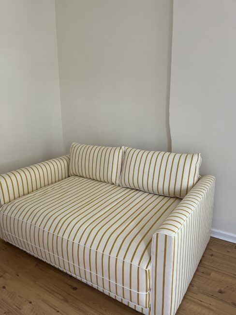 A striped yellow and white sofa with two back cushions sits on a wooden floor against a plain beige wall.