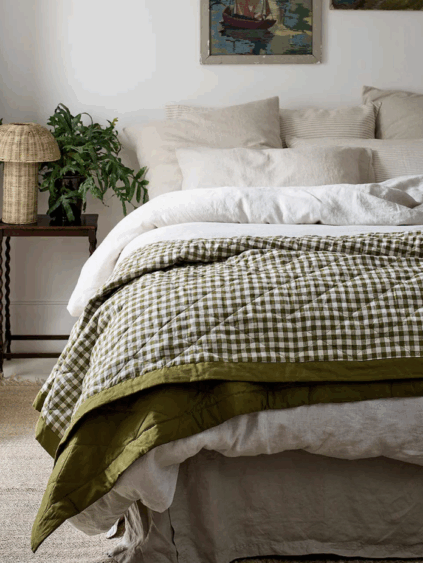 A neatly made bed with light-colored pillows and sheets, topped with a green and white checkered quilt; a side table with a plant and lamp is beside the bed.