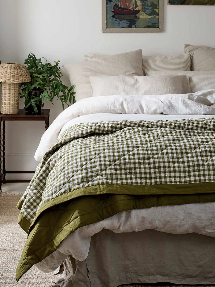 A neatly made bed with light-colored pillows and sheets, topped with a green and white checkered quilt; a side table with a plant and lamp is beside the bed.