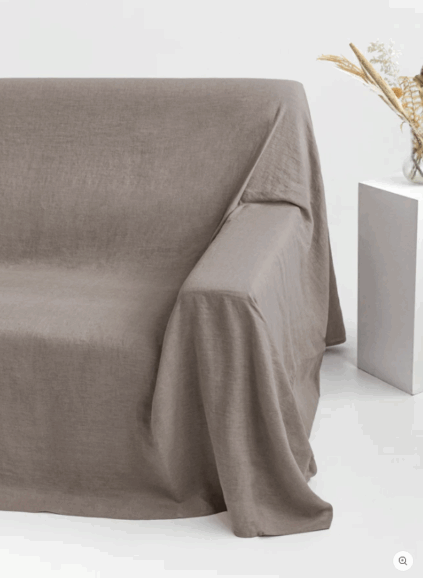 A taupe-colored linen throw covers a sofa next to a white square table with a glass vase holding dried flowers, set against a plain white background.