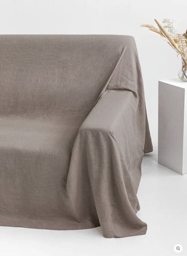 A taupe-colored linen throw covers a sofa next to a white square table with a glass vase holding dried flowers, set against a plain white background.