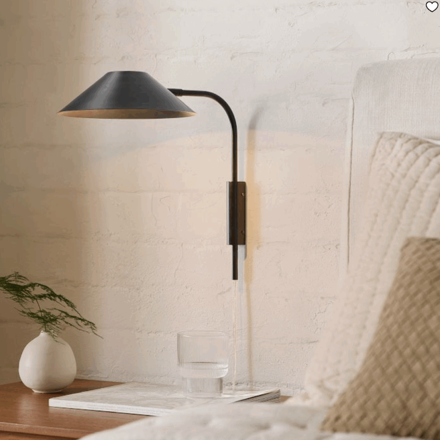 A black wall-mounted lamp is next to a bed with a beige quilt. On the wooden bedside table are a glass of water and a small vase with green foliage.